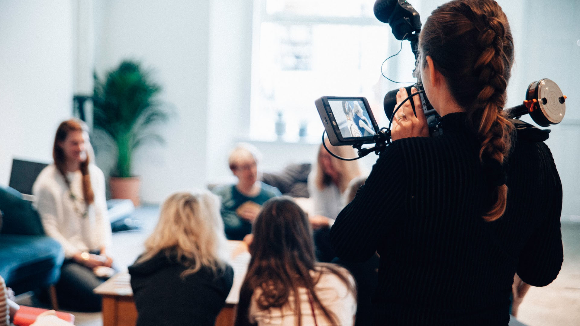 A photograph taken behind a woman holding a camera as she films a group of people seated and talking around a table in a bright room